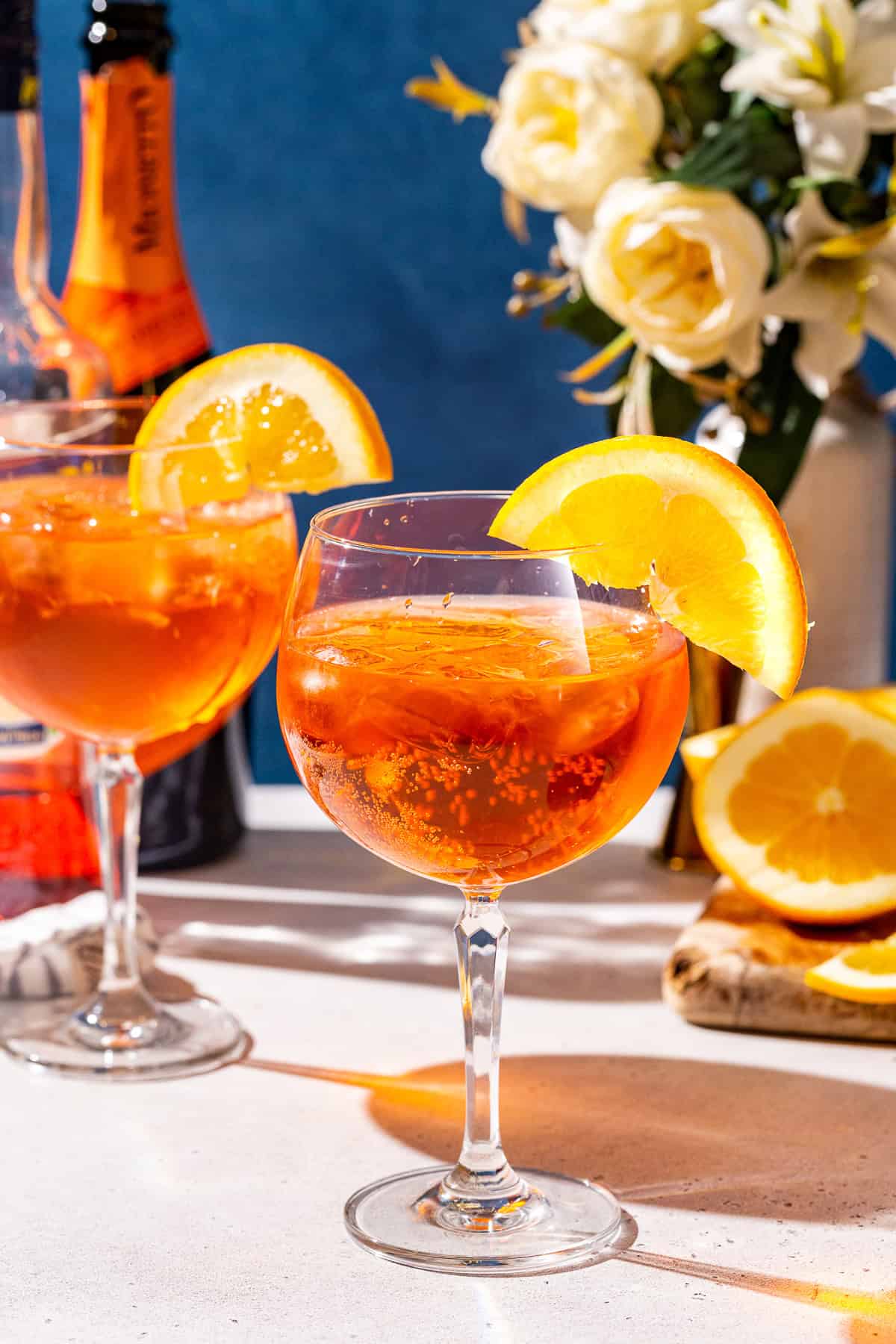 Two glasses of Aperol Spritz with an orange slice garnish on a countertop. Ingredients and bar tools are in the background along with a vase of flowers.