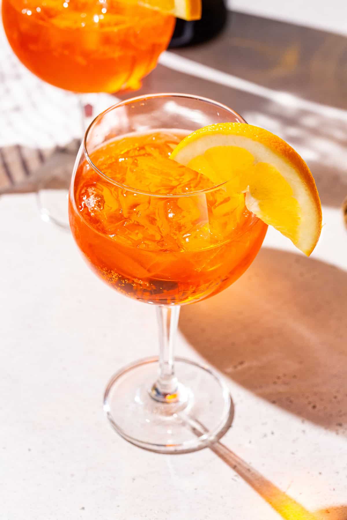 Overhead view of an Aperol Spritz cocktail in a large stemmed cocktail glass. The drink is dark orange in color with an orange slice on the rim as a garnish. Another of the same drink is in the background.