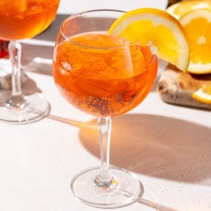 Aperol Spritz cocktail on a countertop with an orange slice garnish. Another spritz is in the background along with a cut orange.