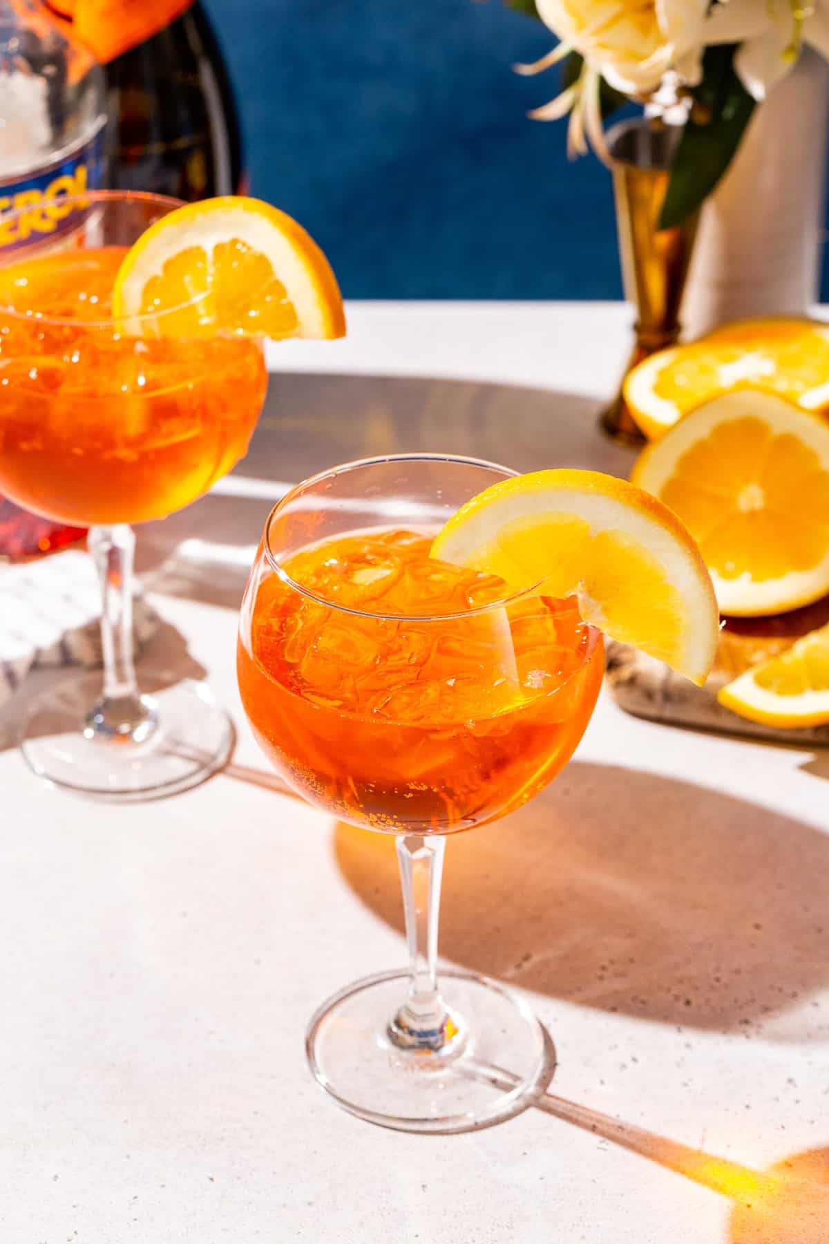 Two Aperol Spritz cocktails on a countertop. The drinks are garnished with orange slices and there are bar tools and ingredients in the background.