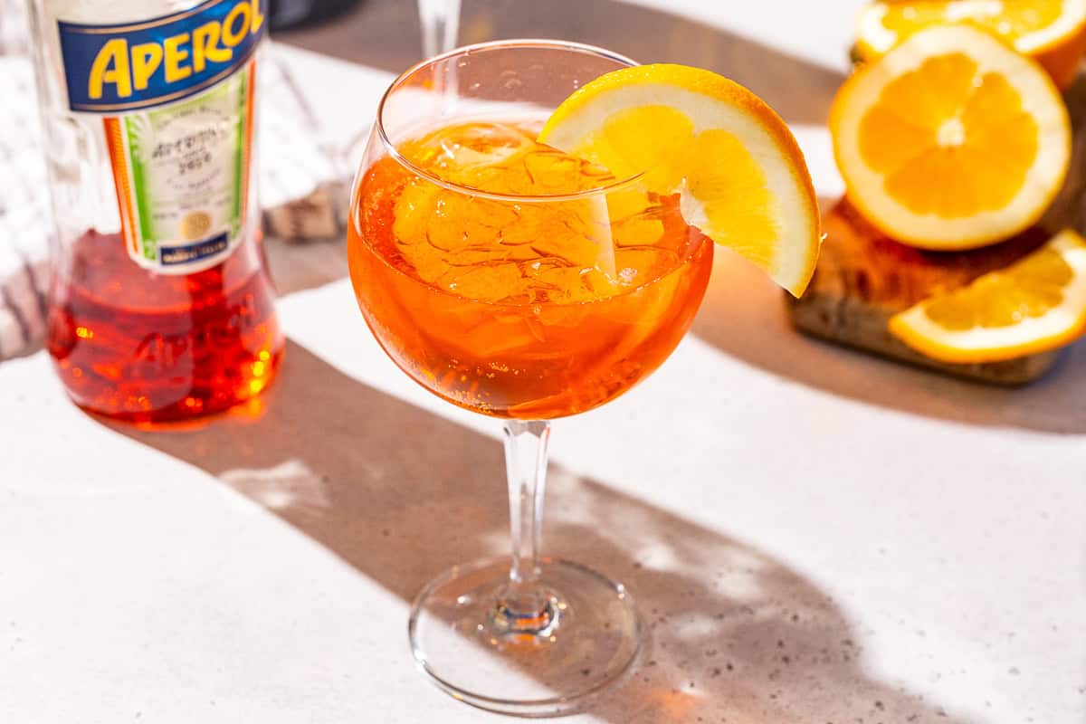 An Aperol Spritz cocktail on a countertop in a stemmed spritz glass. A bottle of Aperol is off to the left.