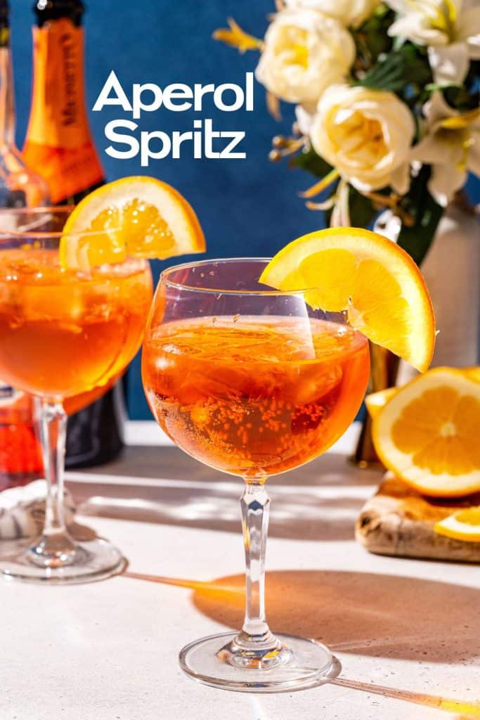 Two glasses of Aperol Spritz with an orange slice garnish on a countertop. Ingredients and bar tools are in the background along with a vase of flowers. Text overlay says “Aperol Spritz”.