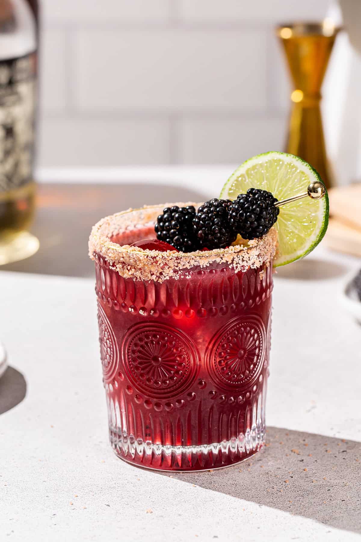 Side view of a Blackberry Margarita tequila cocktail on a countertop. It is garnished with blackberries and a lime slice. There are some ingredients and bar tools in the background.