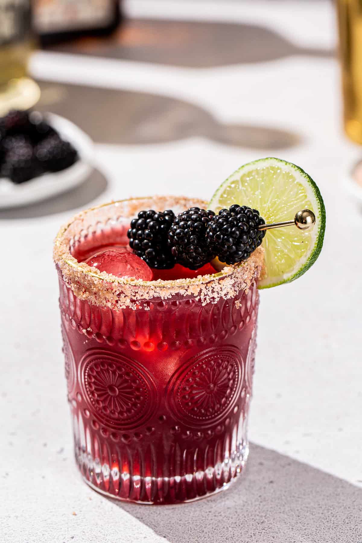 Blackberry Margarita on a countertop with blackberries in a dish in the background. The drink is in an embossed cocktail glass with a salted rim, and garnished with fresh blackberries and a lime slice.