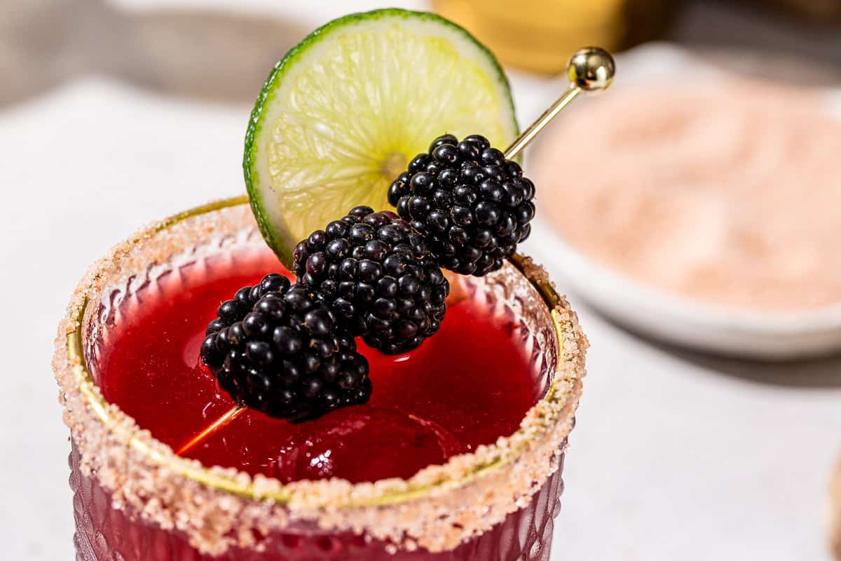 Close up of a Blackberry Margarita with a salt rim. The drink is garnished with a lime slice and three blackberries.