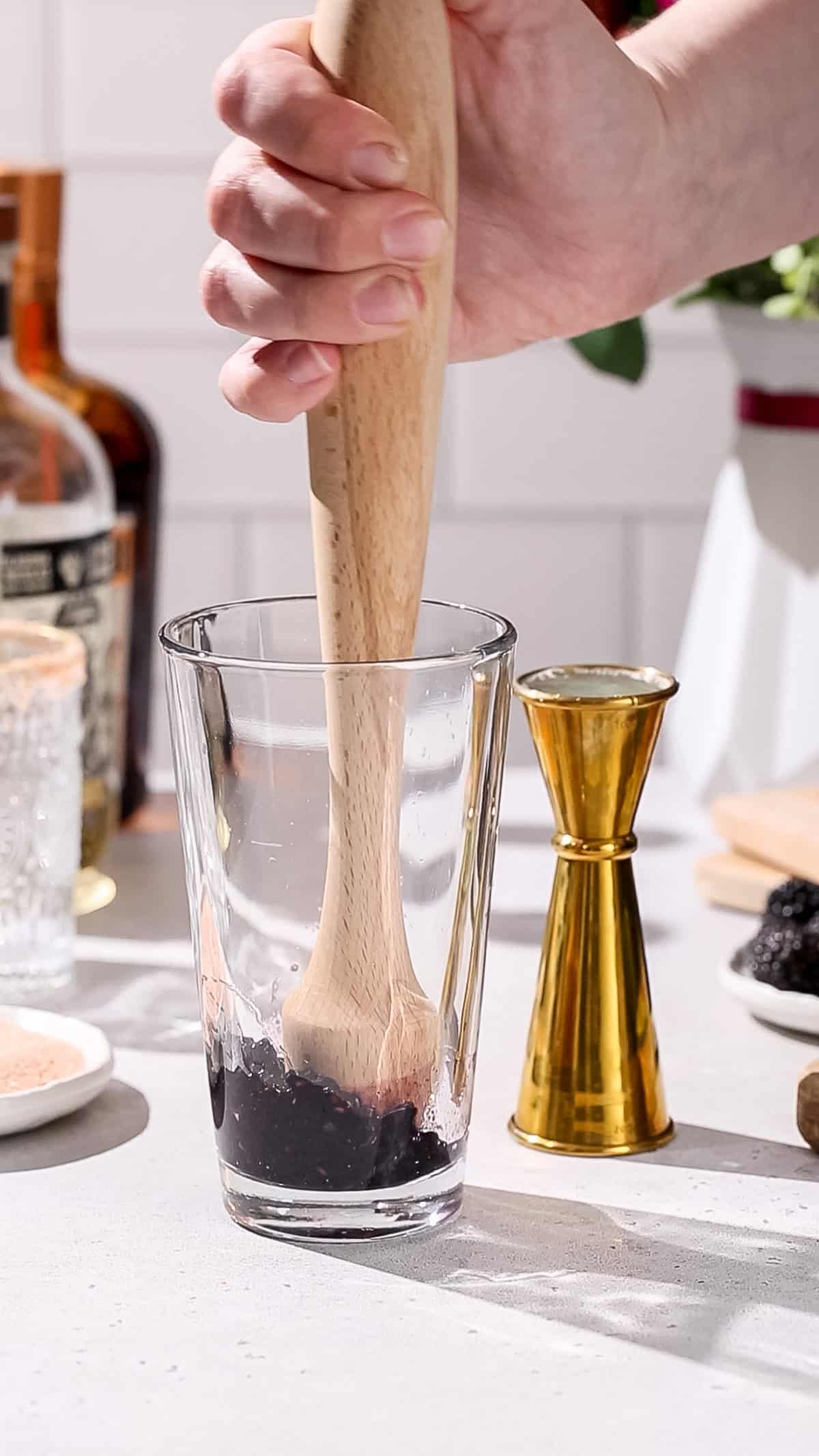 Hand using a wooden muddler to smash blackberries in a cocktail shaker.