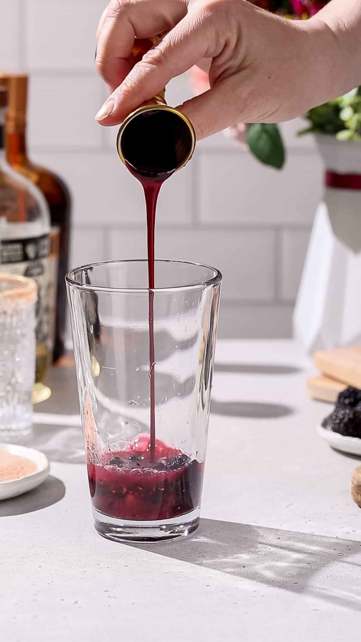 Hand pouring blackberry syrup into a cocktail shaker.