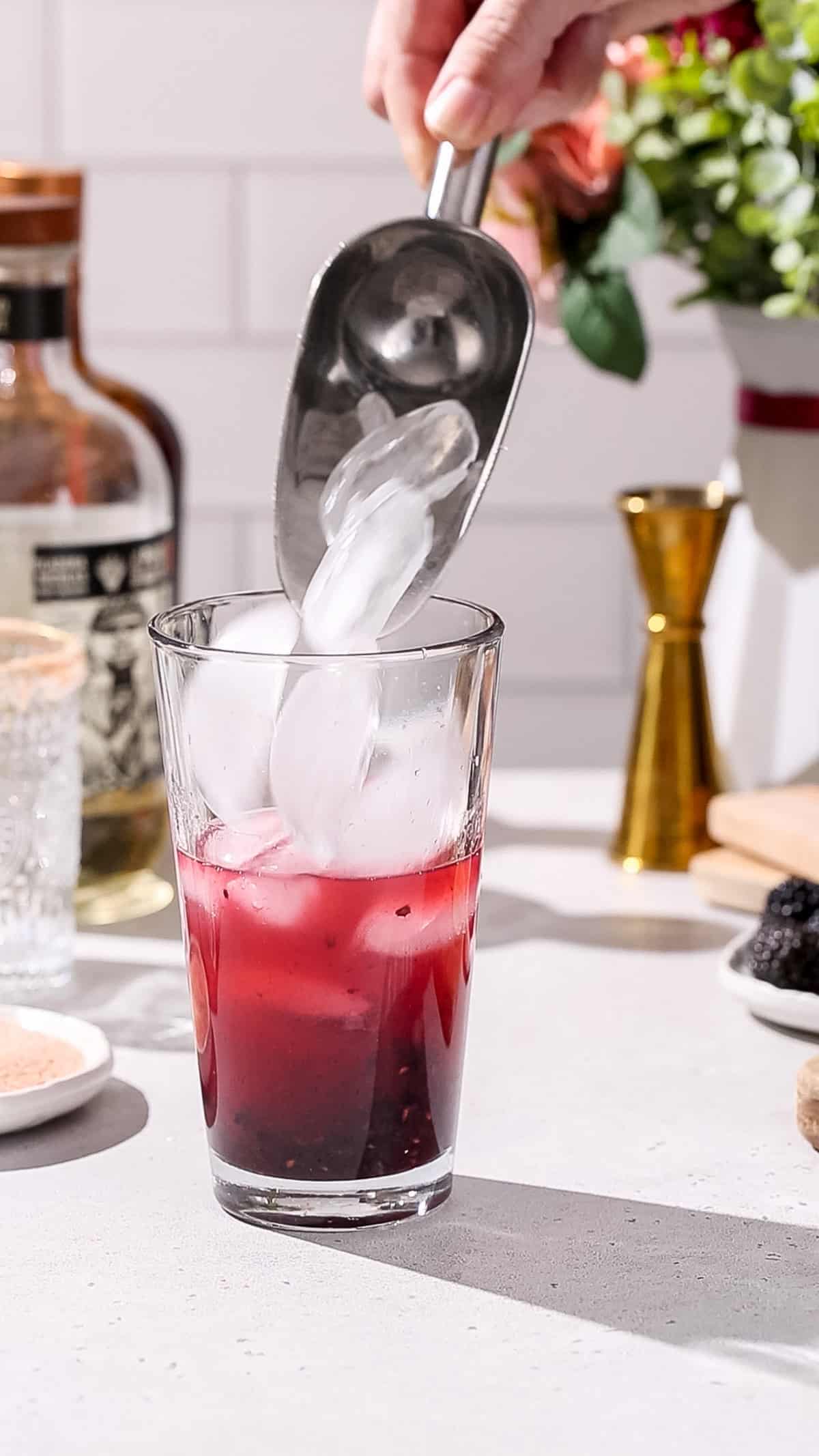 Hand using an ice scoop to add ice to a glass cocktail shaker.