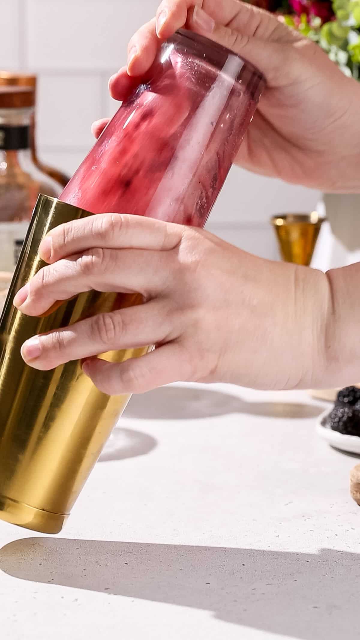 Hands shaking a cocktail shaker filled with a dark red liquid.