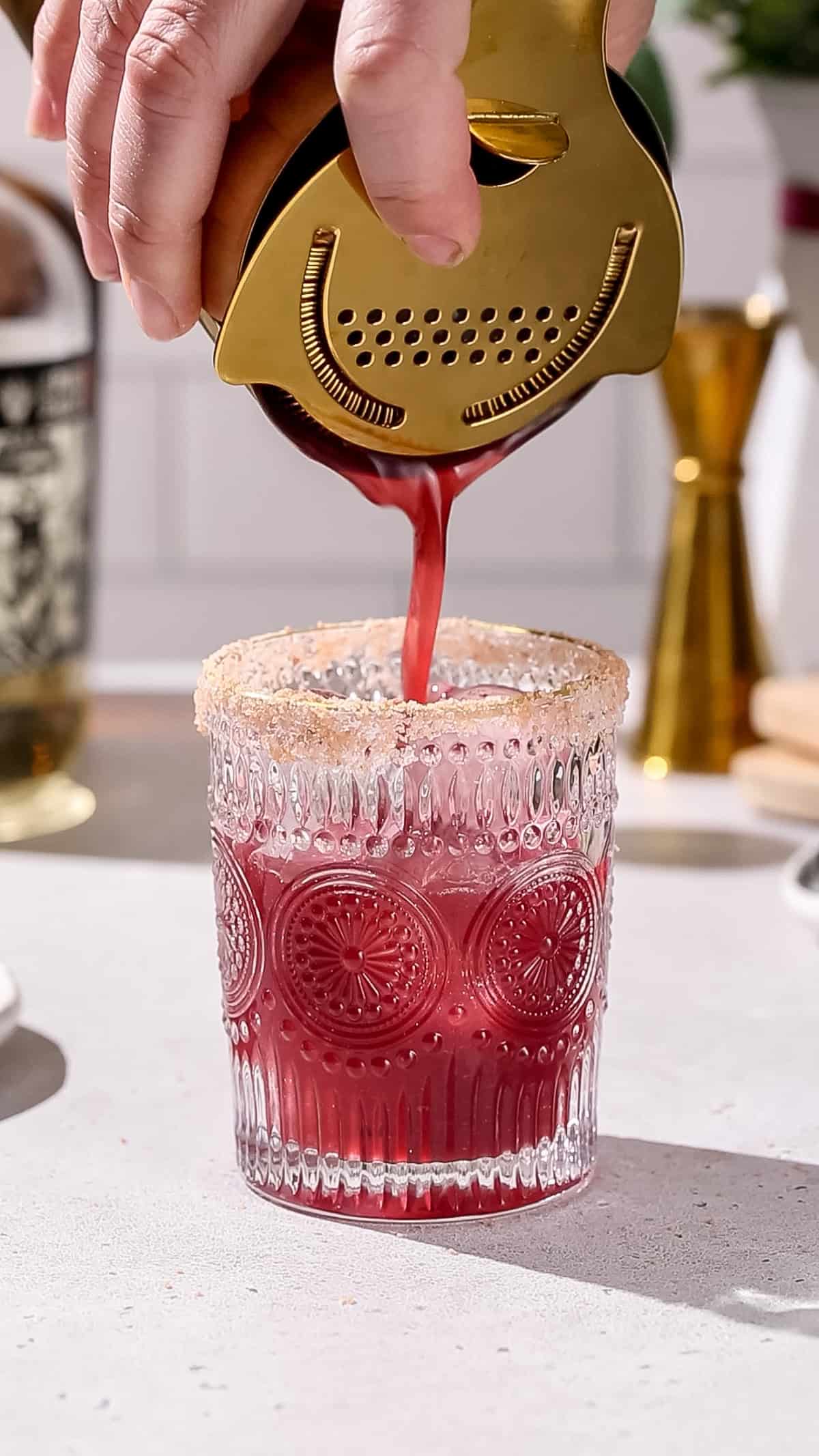 Hand straining a dark red liquid into an embossed cocktail glass. The glass is filled with ice and has salt on the rim.