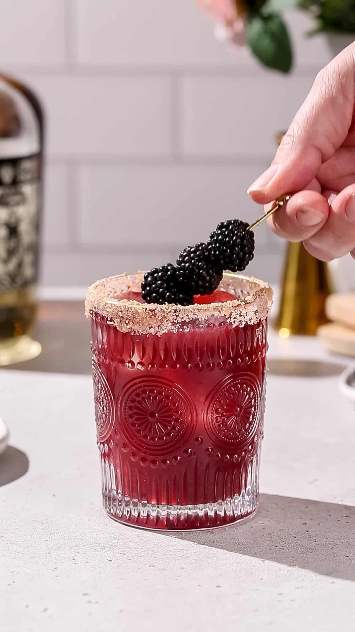 Hand adding a cocktail pick with blackberries on it to garnish a red cocktail.