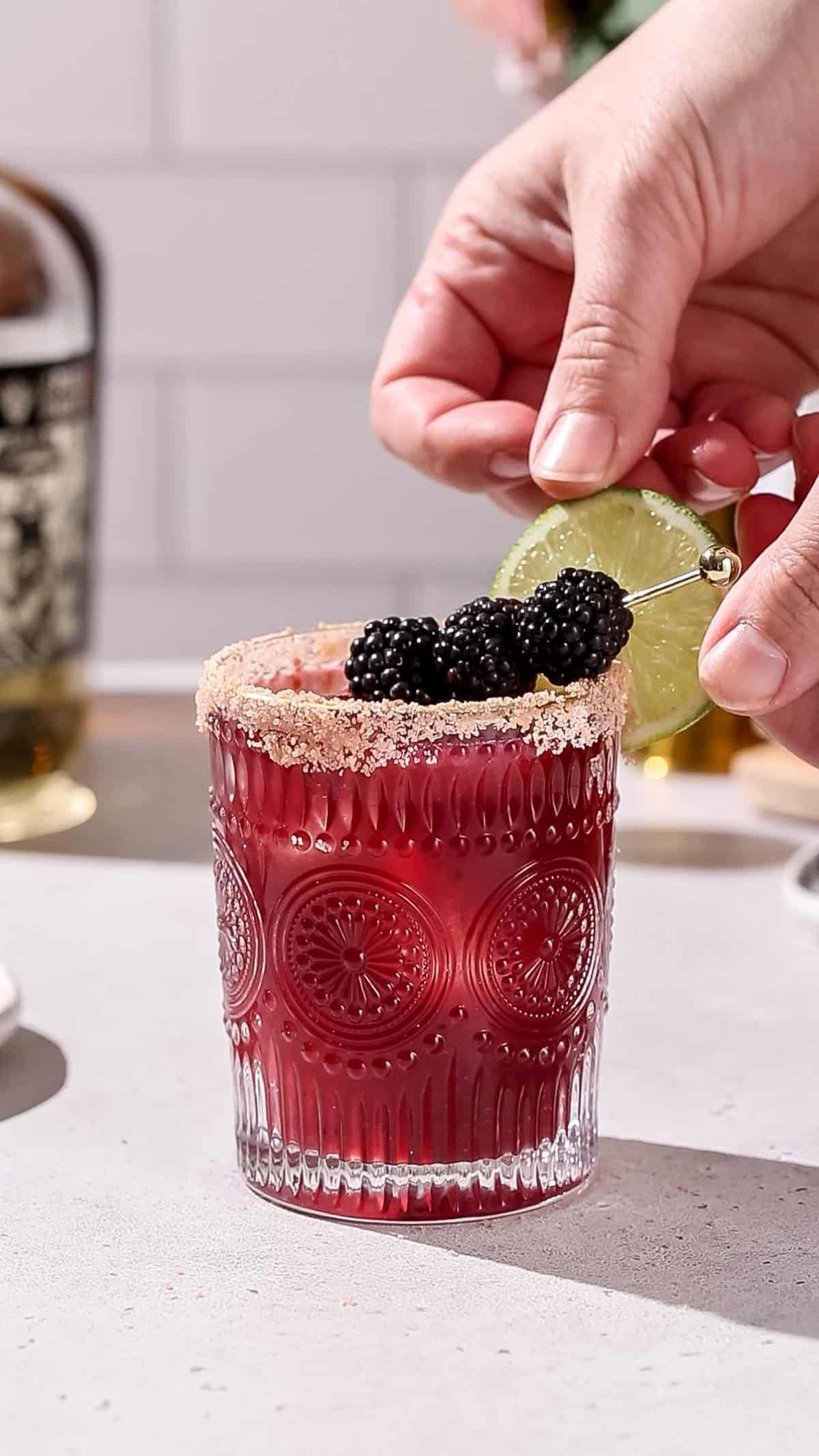 Hand adding a lime slice to the rim of a blackberry margarita.