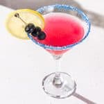 Martini glass with a Blueberry Lemon Drop cocktail. The drink is red with blue sugar rim and garnished with a lemon slice and blueberries.