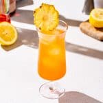 Tropical Whiskey Punch cocktail in a tall hurricane glass. The drink is orange and filled with small ice spheres and has a dried pineapple garnish on top. In the background are ingredients and bar tools used to make the drink.