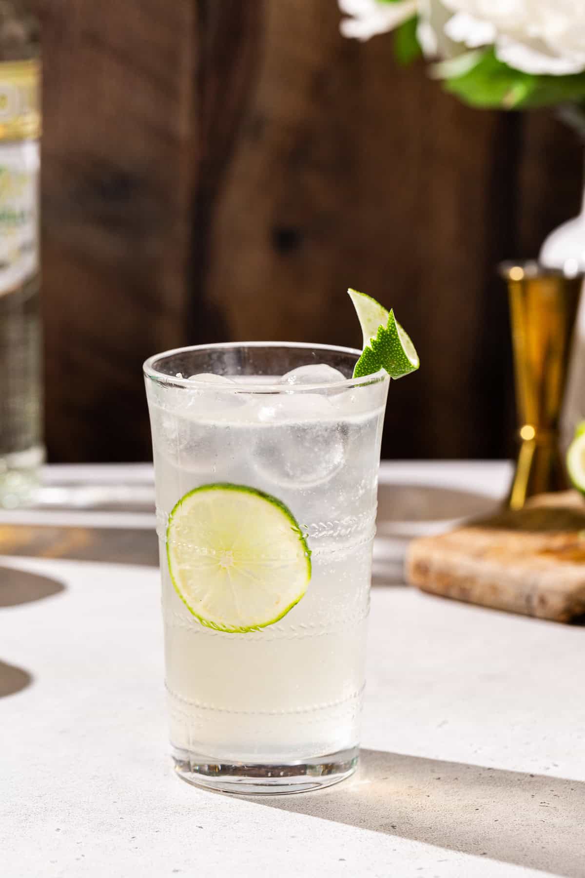 Vanilla Lime Vodka Refresher cocktail on a countertop. A jigger and small cutting board are seen in the background along with some liquor bottles.