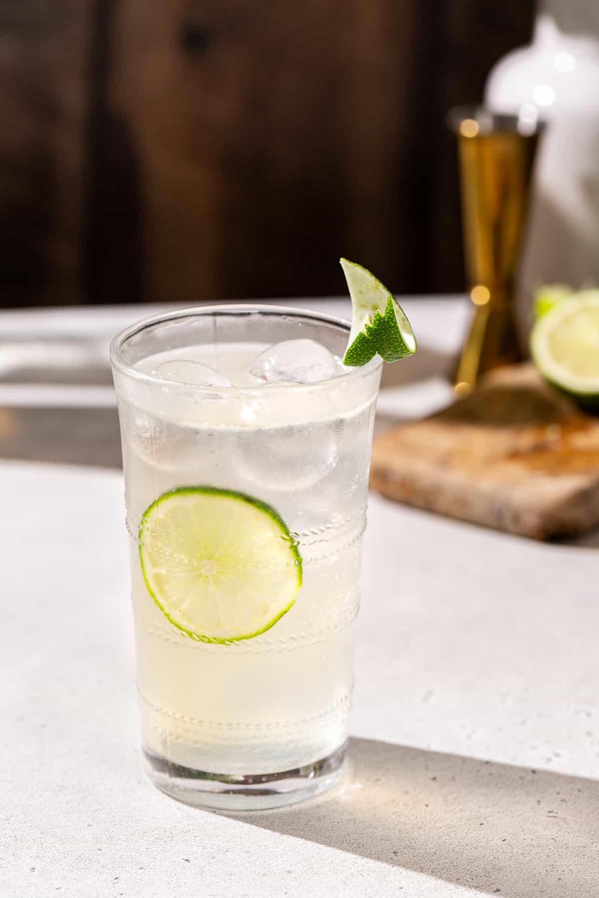 Vanilla Lime Vodka Refresher cocktail with a lime wheel on the inside of the glass and a lime peel garnish on the rim of the glass. A cutting board with a cut lime and a jigger are in the background.