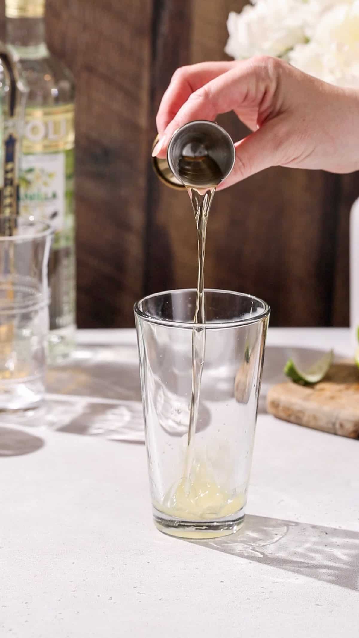 Hand pouring elderflower liqueur into a cocktail shaker.