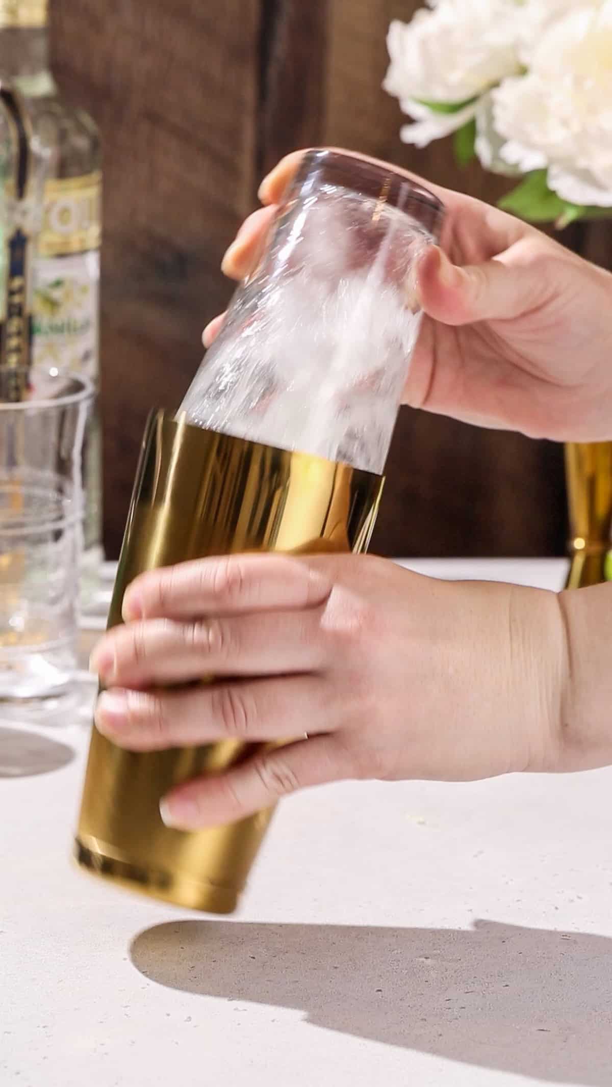 Hands shaking a cocktail shaker filled with ice and clear liquid.