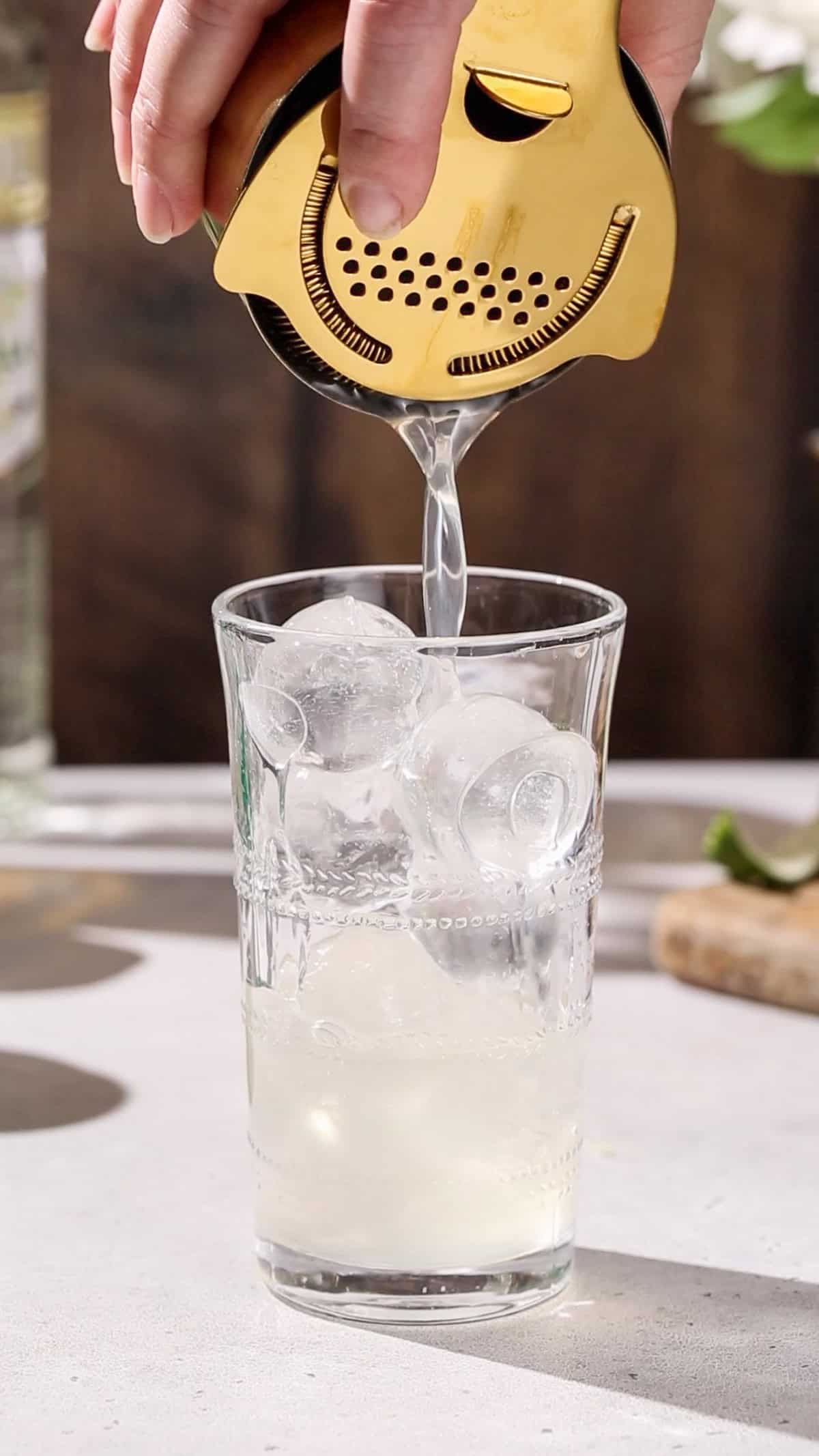 Hand using a cocktail strainer to strain the liquid into a glass filled with spherical ice cubes.