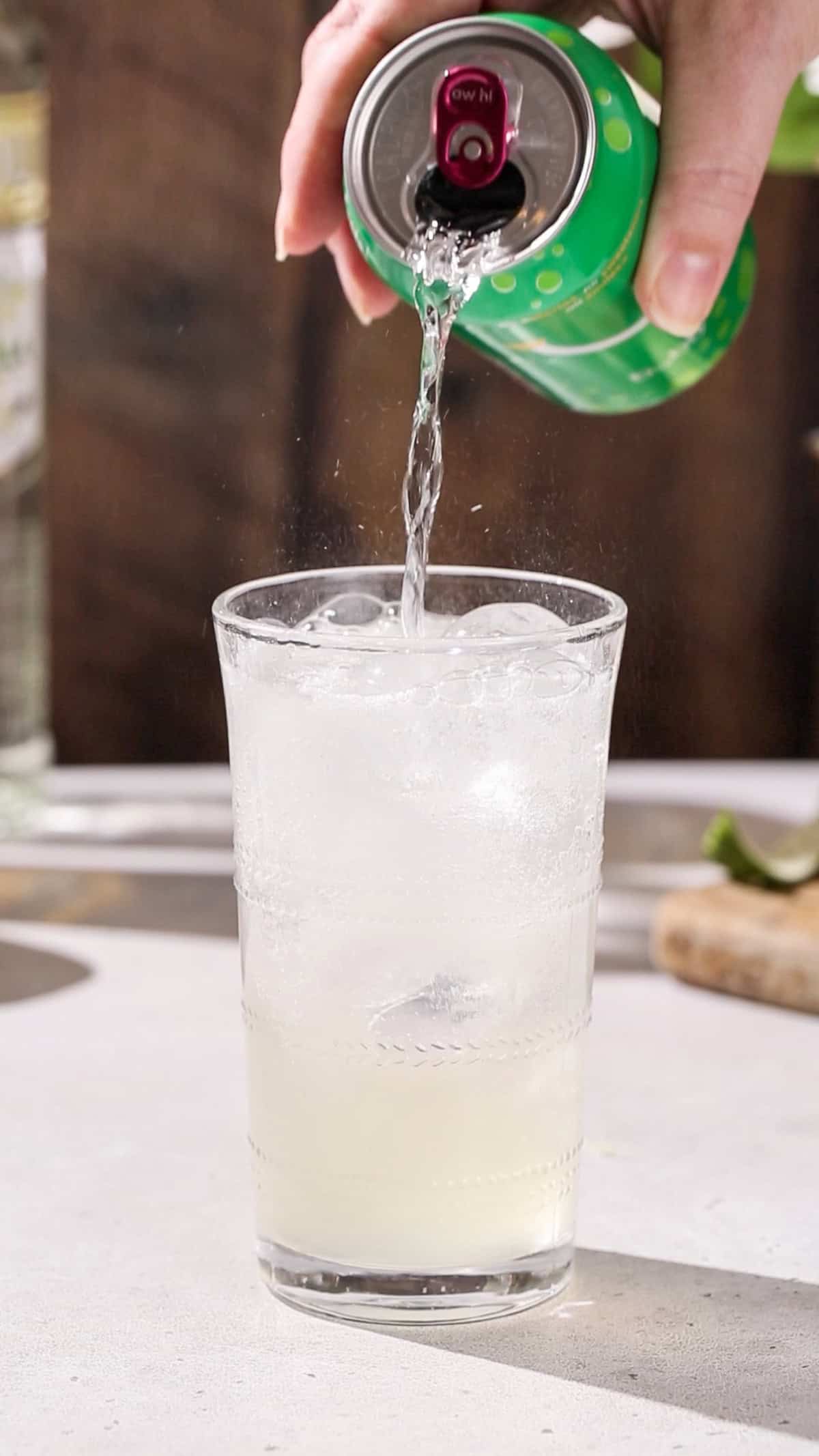 Hand pouring lime seltzer into a cocktail glass.