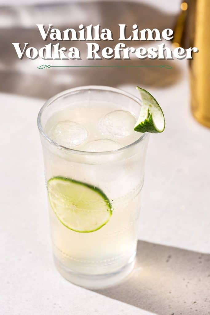 Slightly overhead view of a cocktail glass filled with liquid and spherical ice cubes. There is a lime slice on the inside of the glass, and a carved piece of lime peel garnishes the drink on the rim.