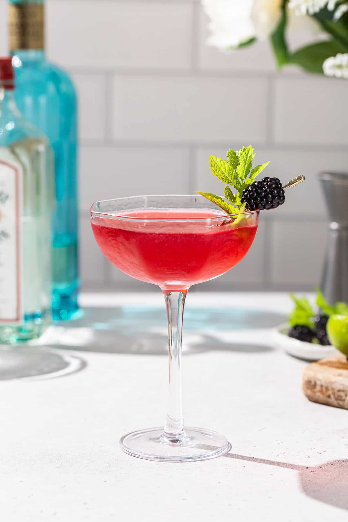 Side view of a Blackberry Bergamot gimlet in a stemmed coupe glass. The drink is red and it has a blackberry and fresh mint garnish. In the background are some bar tools and ingredients to make the drink.