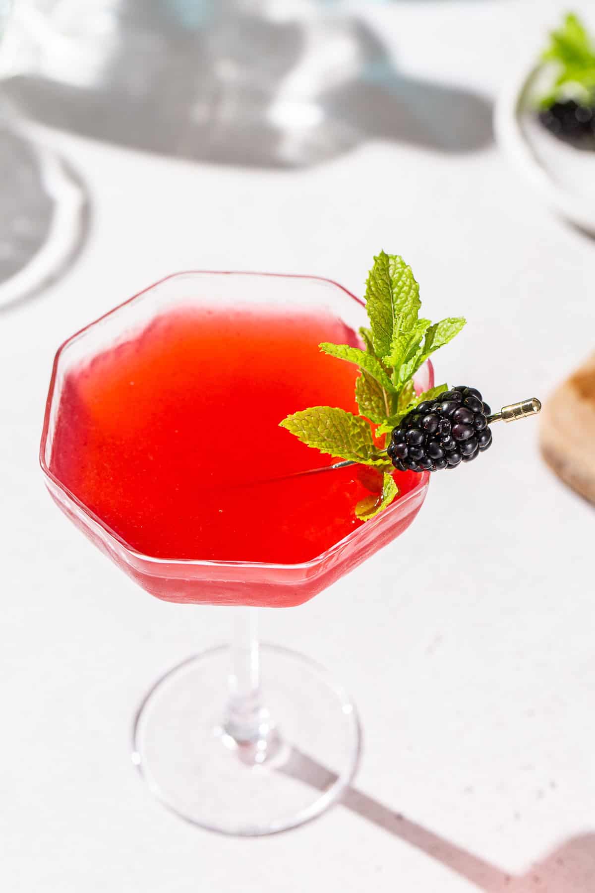 Overhead view of a red colored Blackberry Bergamot gin gimlet in a coupe glass. There is fresh mint and blackberry garnishing the drink.