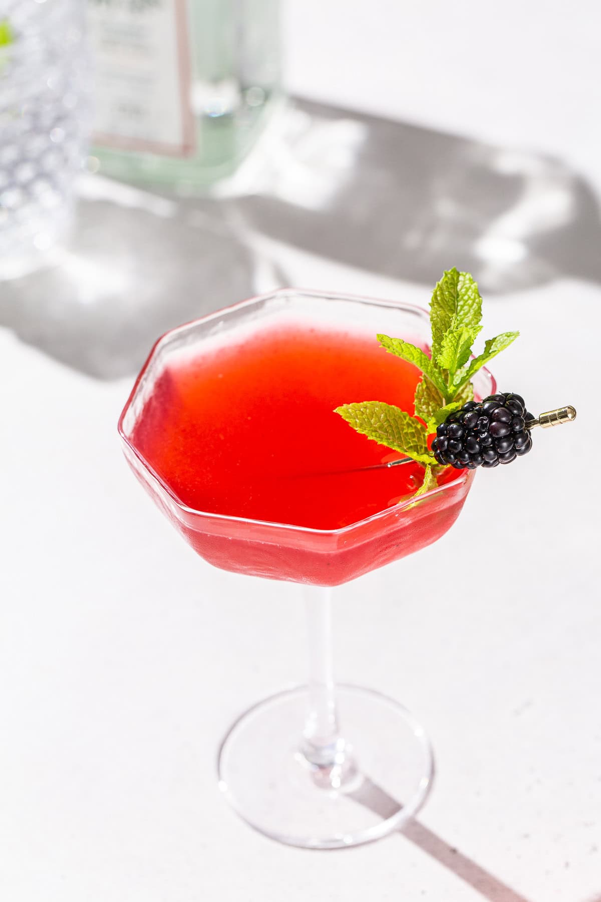 Blackberry Bergamot gin gimlet in a coupe glass on a white countertop. The drink is bright red and garnished with mint and a blackberry.