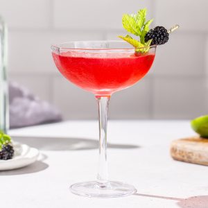 Side view of a Blackberry Bergamot gin gimlet in a coupe glass. The drink has a fresh mint and blackberry garnish, and some bar tools and ingredients can be seen in the background.