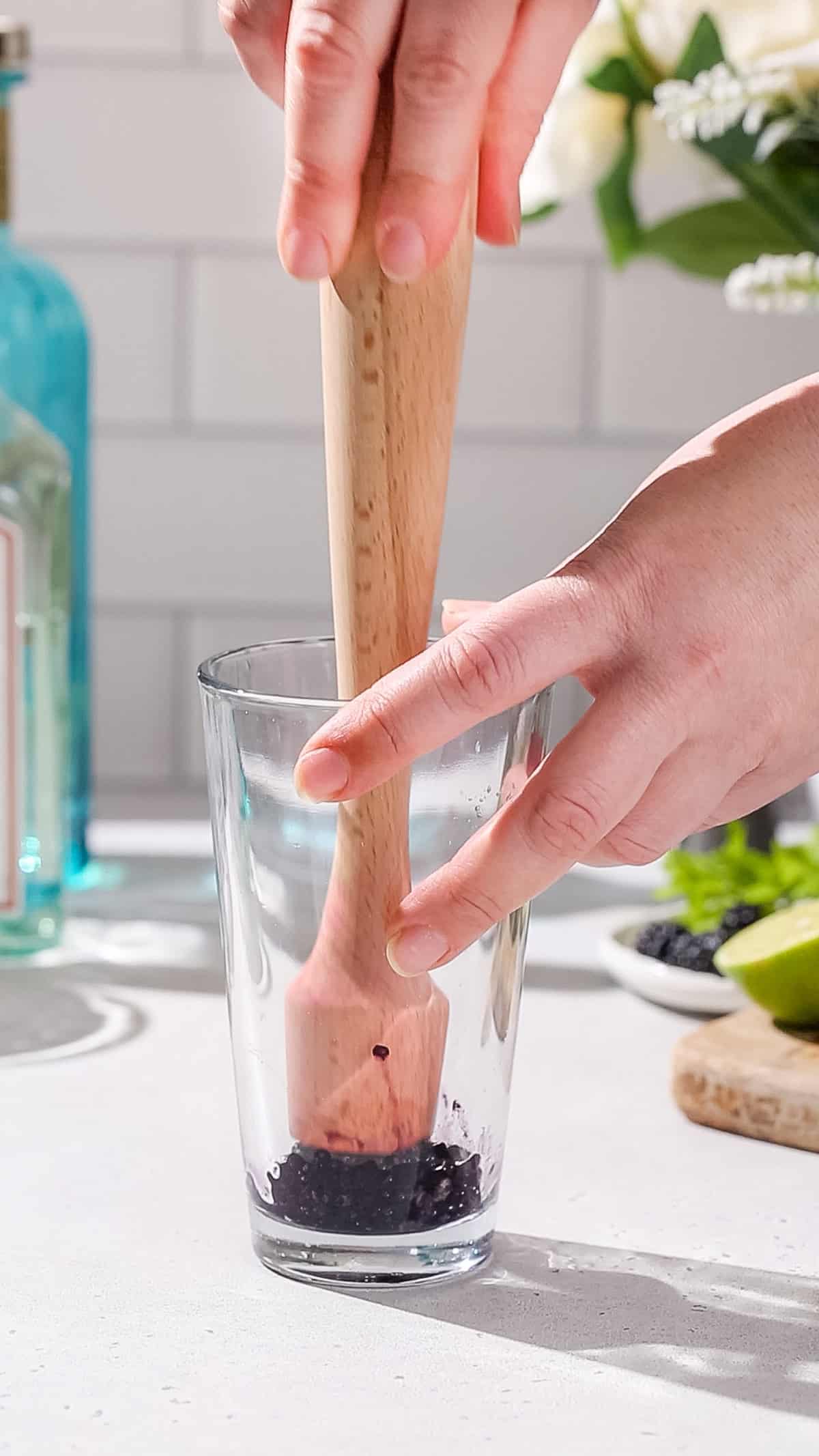 Hand using a wooden muddler to smash blackberries in a cocktail shaker.