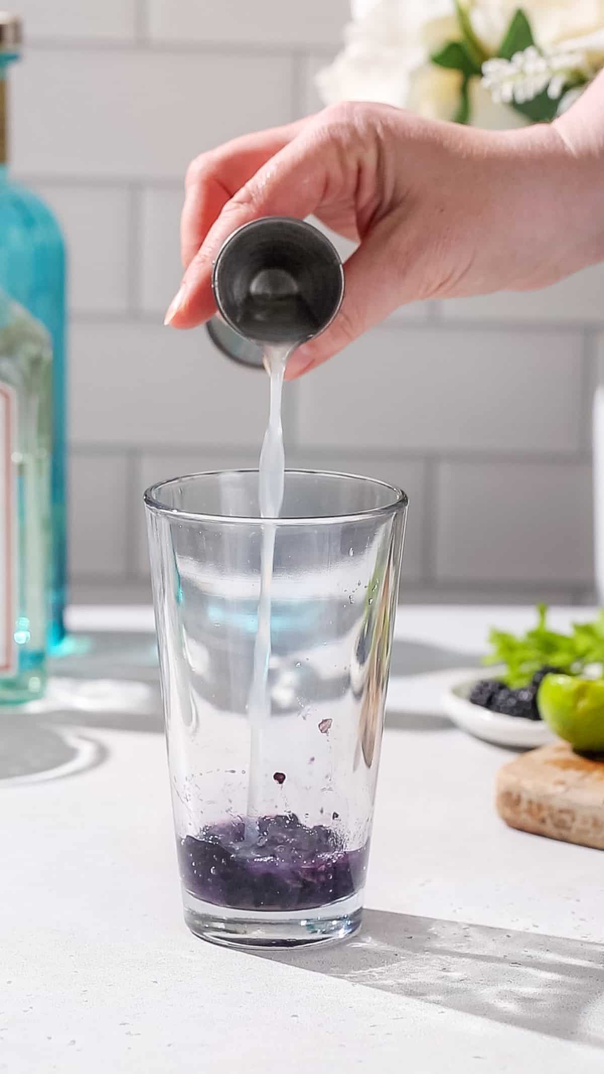 Hand pouring fresh lime juice into a cocktail shaker with blackberries in it.