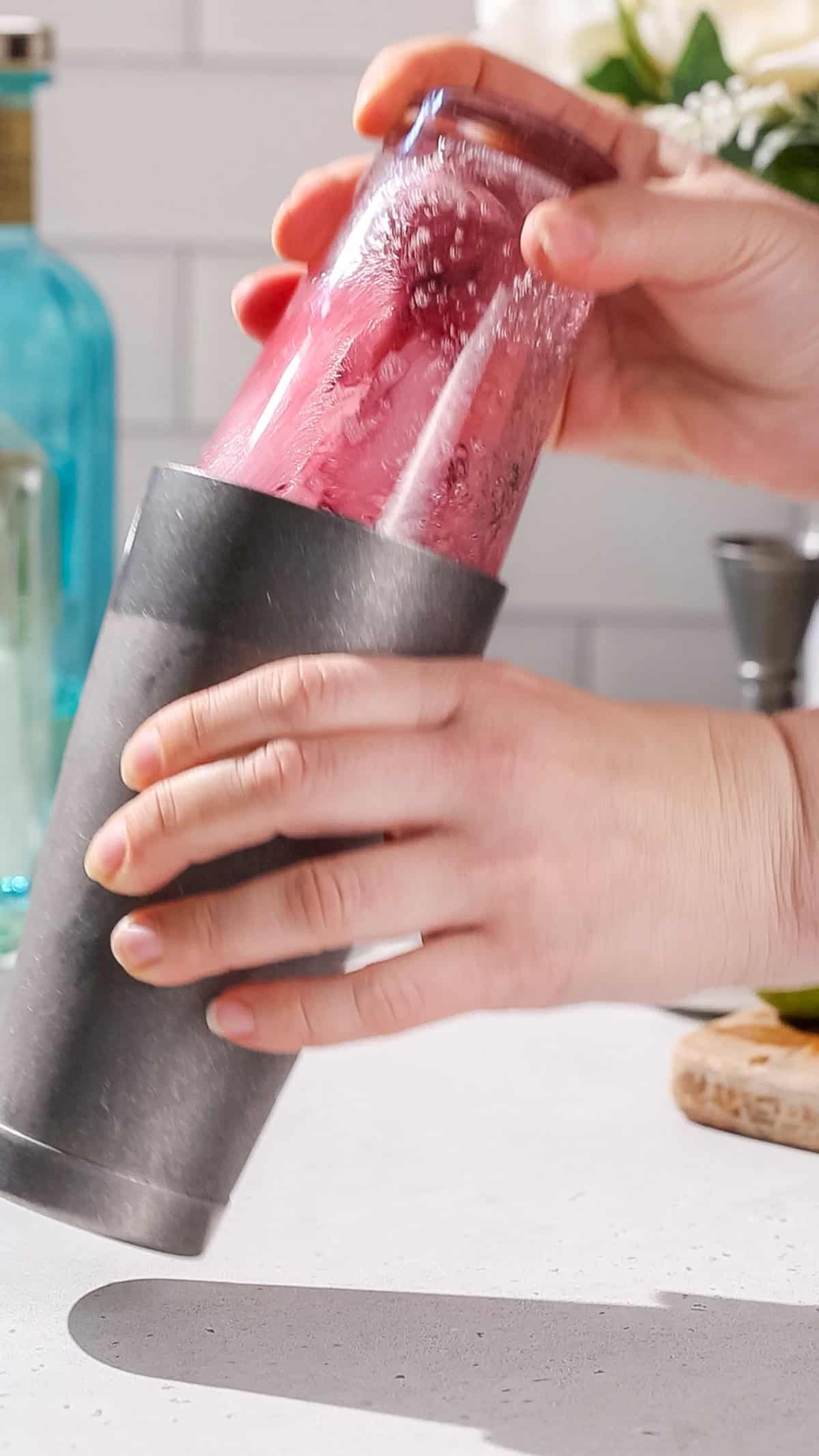 Hands shaking up a cocktail shaker filled with a purplish red mixture.