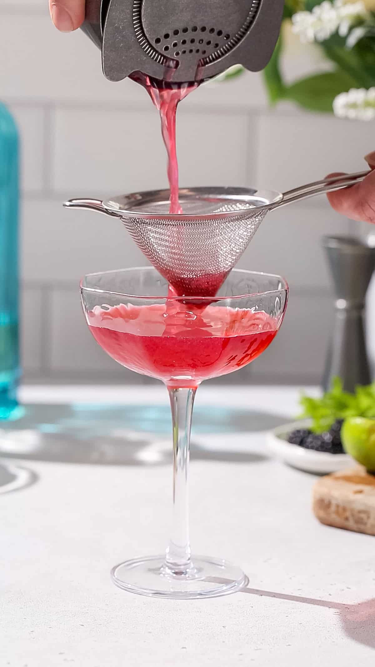 Hand straining a red cocktail into a coupe glass using a regular strainer and a fine mesh strainer.