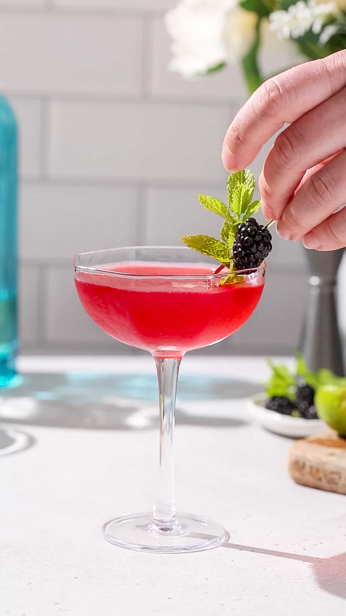 Hand adding a blackberry and mint garnish to a red cocktail.