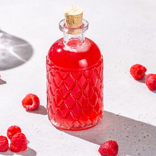 Raspberry simple syrup in a glass bottle with some fresh raspberries next to it on the counter.