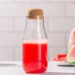 Watermelon juice in a corked bottle on a countertop. Fresh watermelon is in the background.