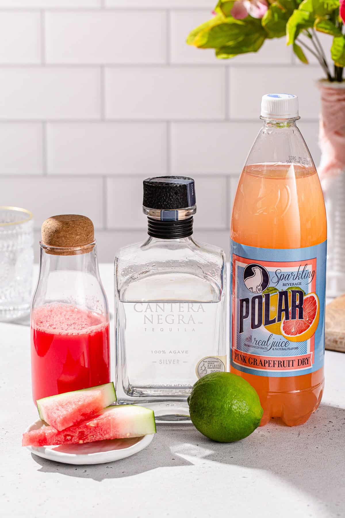 Ingredients to make a Watermelon Paloma cocktail together on a countertop, including grapefruit soda, tequila and watermelon juice.