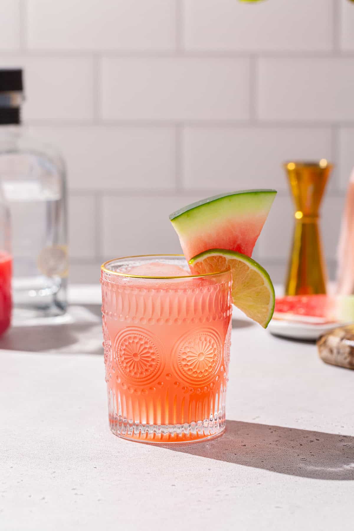 Watermelon Paloma on a countertop with a bottle of tequila, a jigger, watermelon juice and fresh watermelon in the background.