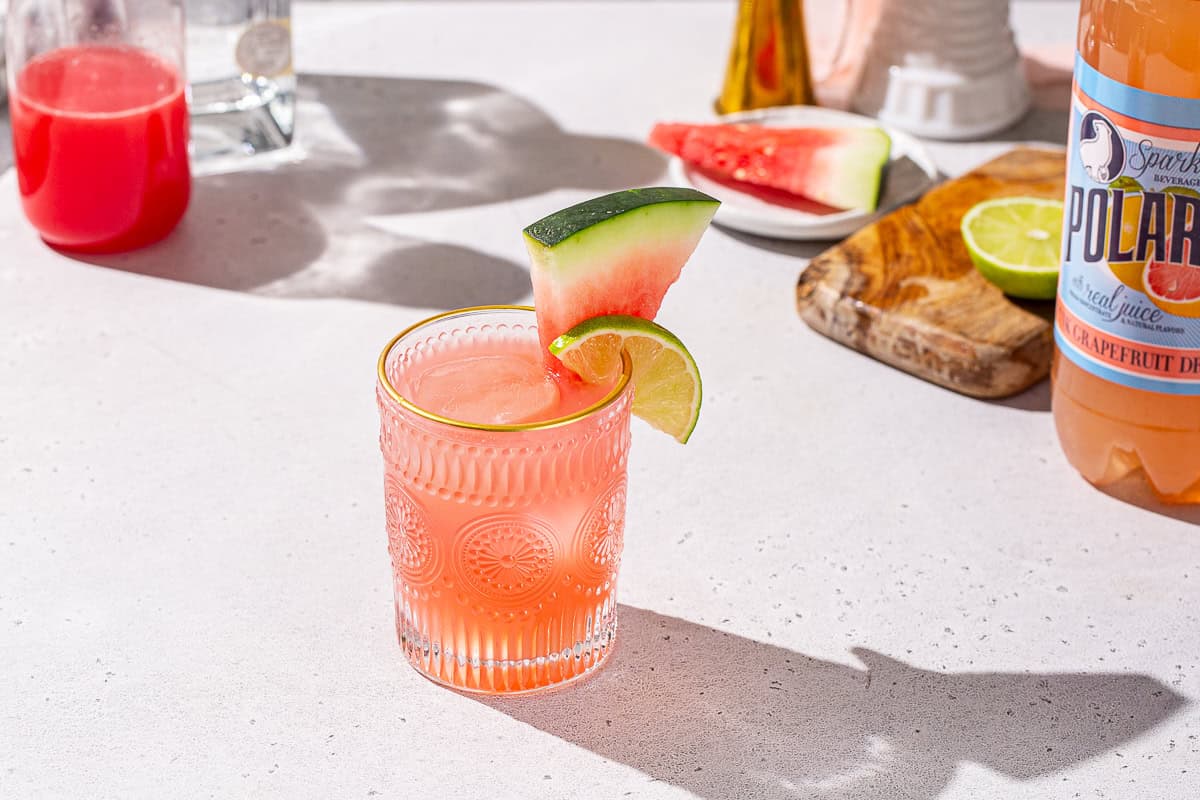 Watermelon Paloma on a countertop with some bar tools and ingredients to make the drink in the background. The drink is garnished with a watermelon wedge and lime slice. A bottle of grapefruit soda is off to the right.