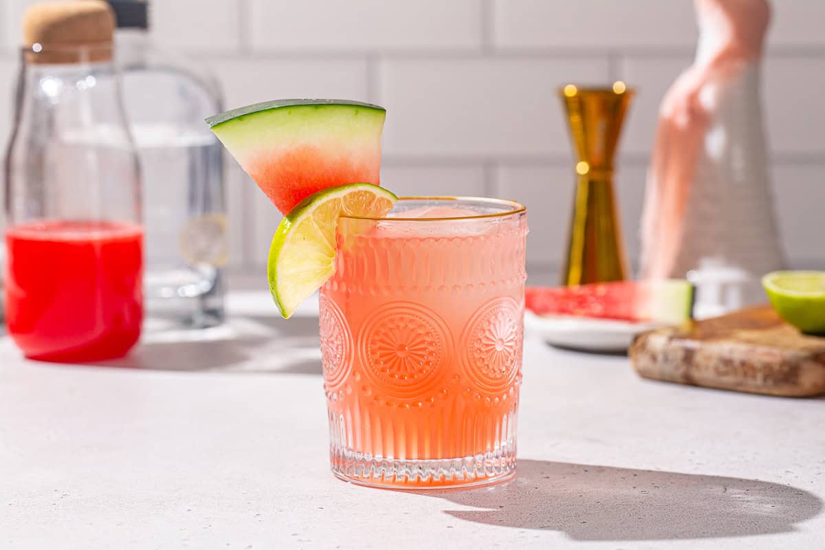 Side view of a Watermelon Paloma cocktail with a jigger, tequila bottle, and watermelon wedges in the background, along with watermelon juice and a cut lime.