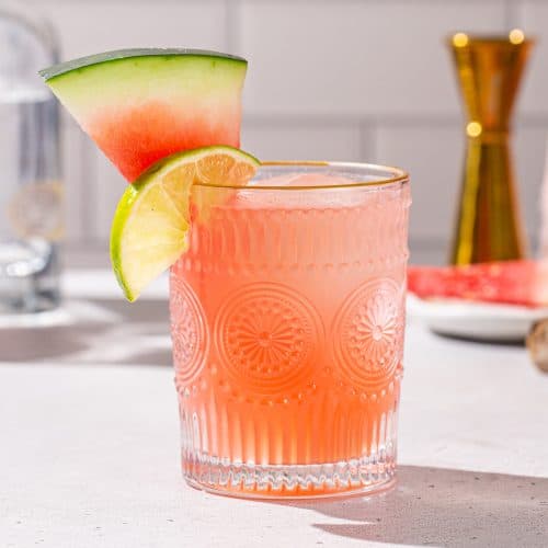 Watermelon Paloma cocktail on a countertop with a watermelon slice and lime slice as garnish.