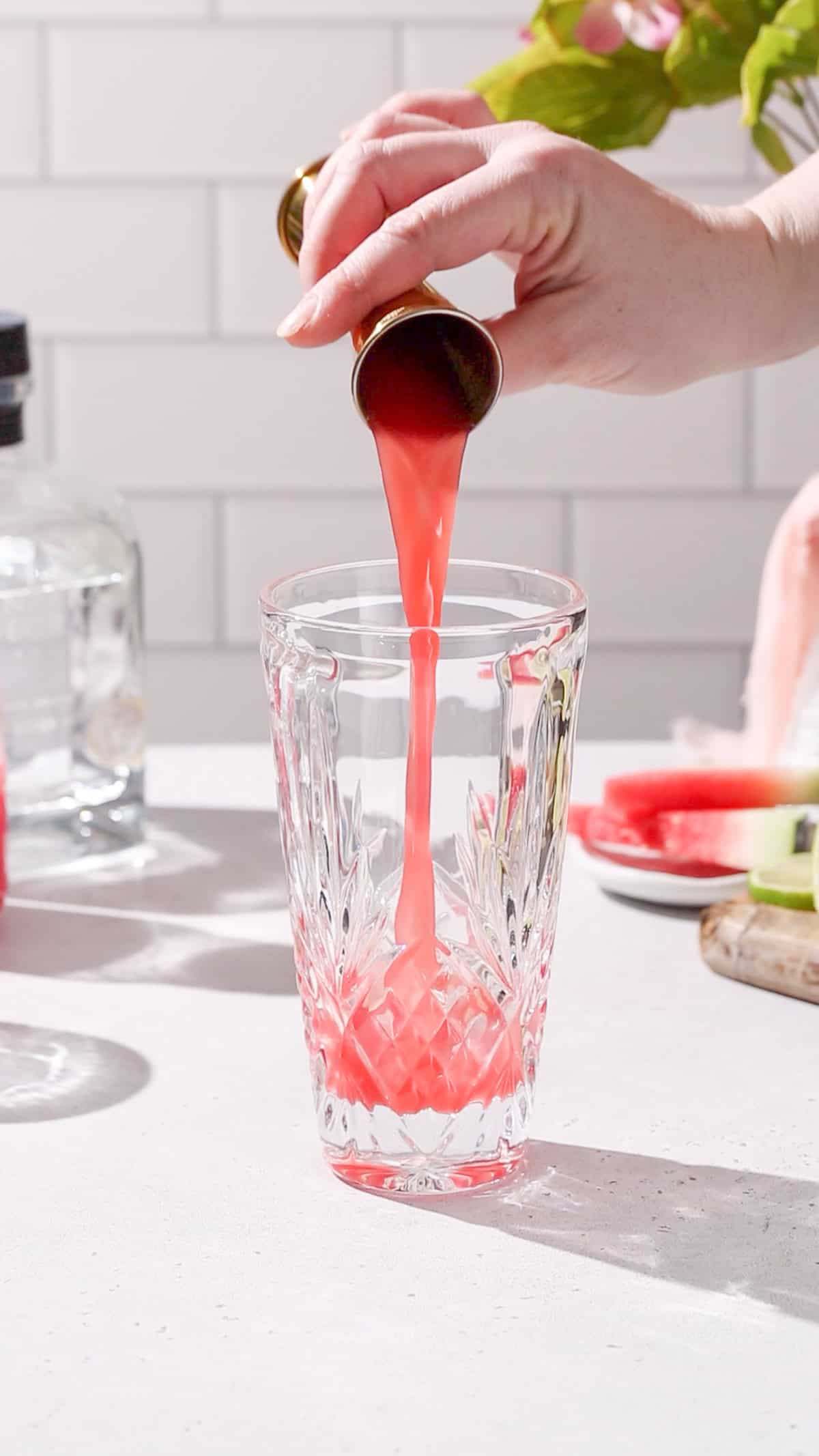 Hand pouring watermelon juice from a jigger into a cocktail shaker.