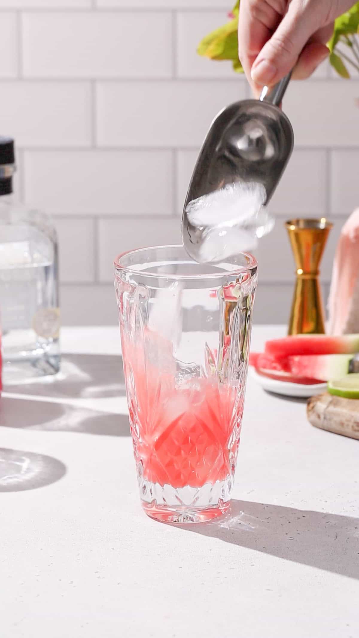 Hand adding ice to a cocktail shaker filled with red liquid.