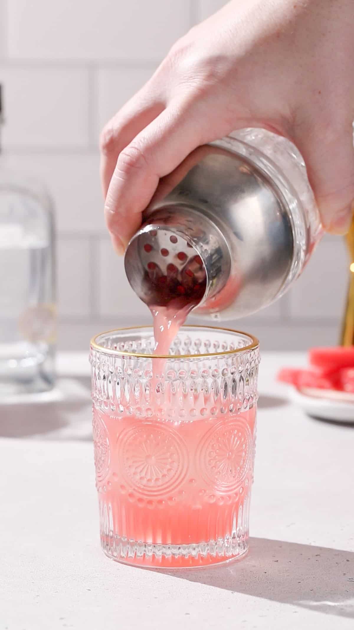 Hand using a cocktail shaker to strain a pink colored drink into a gold-rimmed embossed cocktail glass.