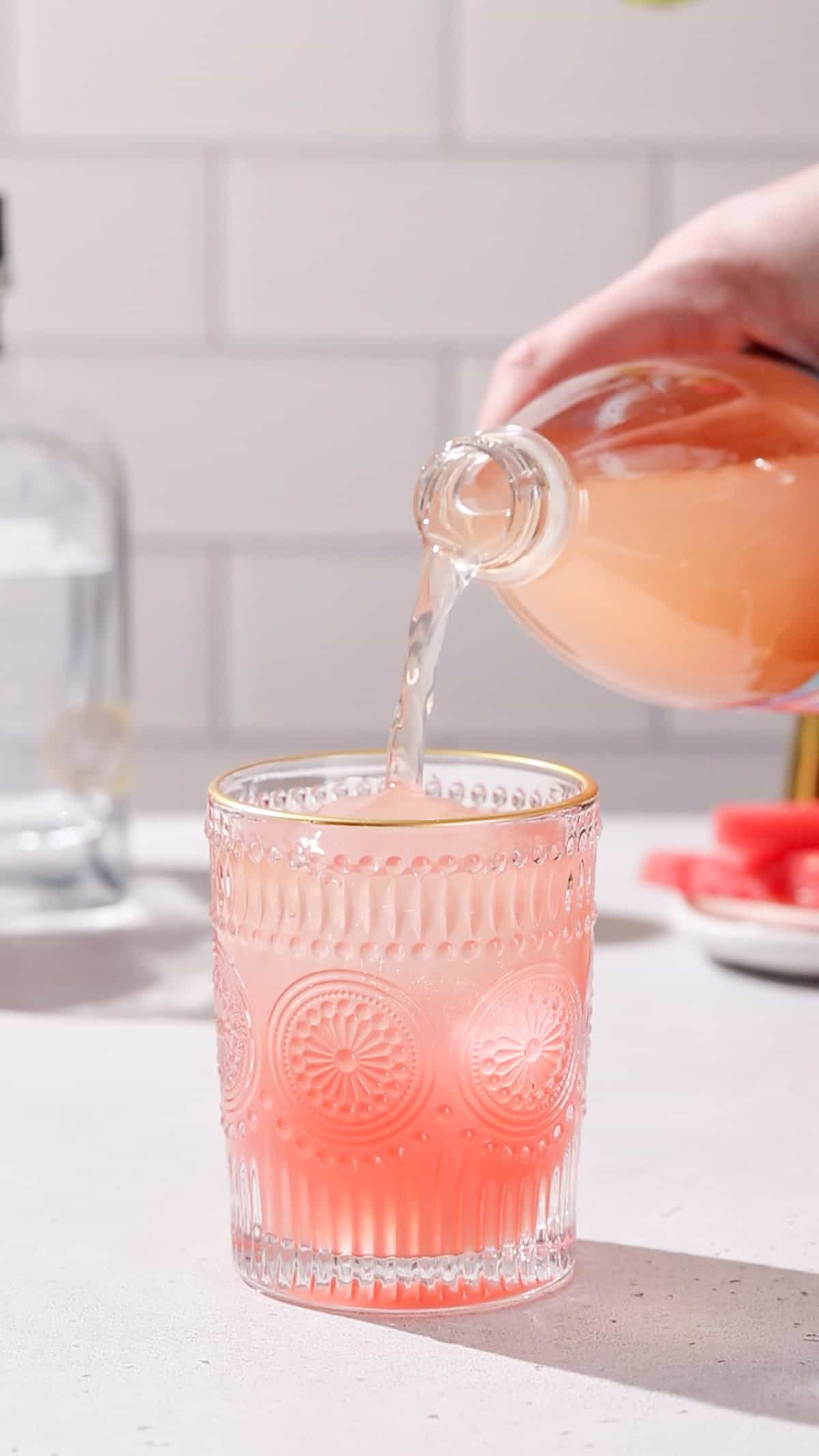 Hand pouring grapefruit soda into a cocktail glass filled with pink liquid.