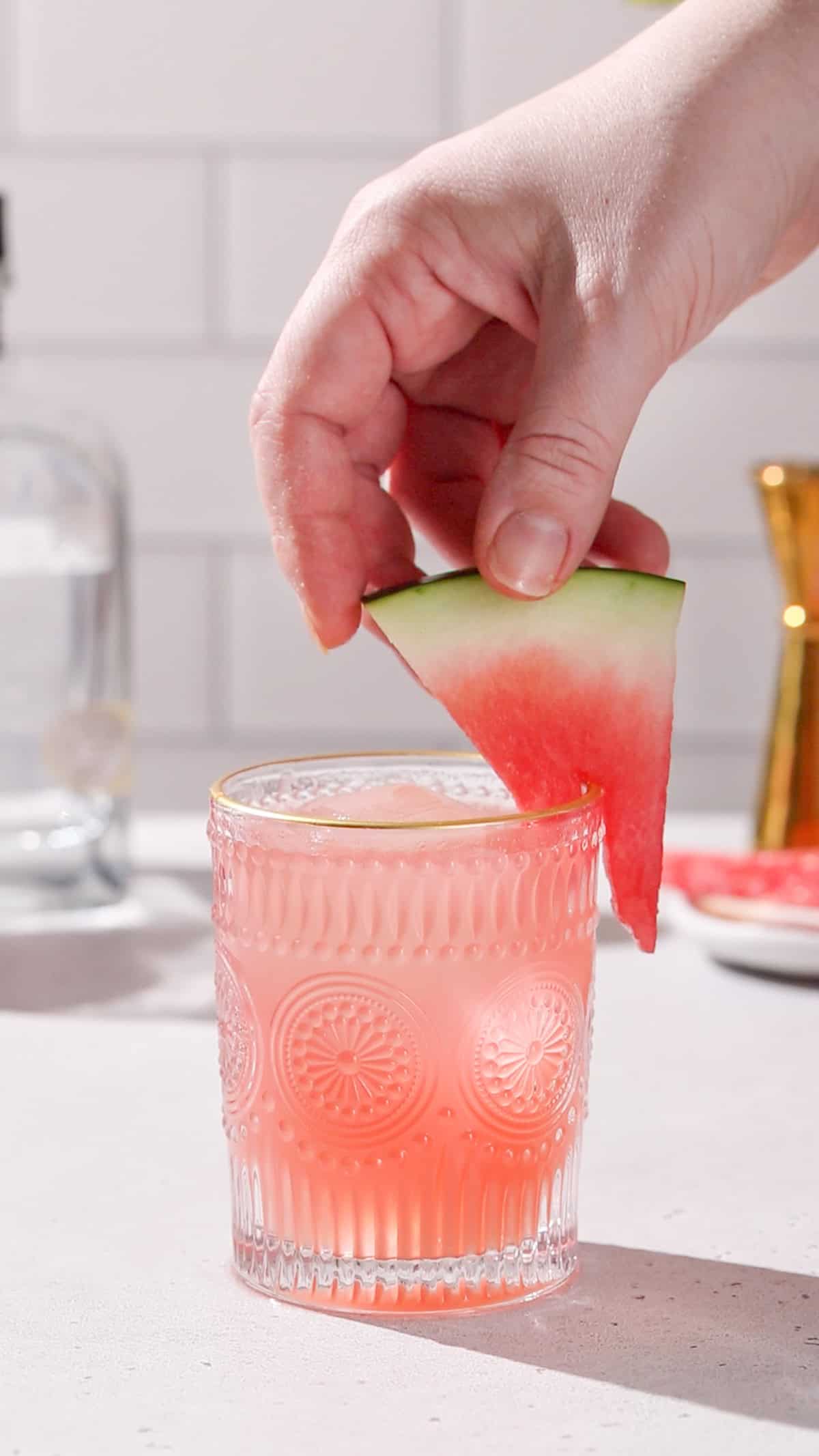 Hand adding a watermelon wedge to the edge of a glass filled with ice and pink liquid.