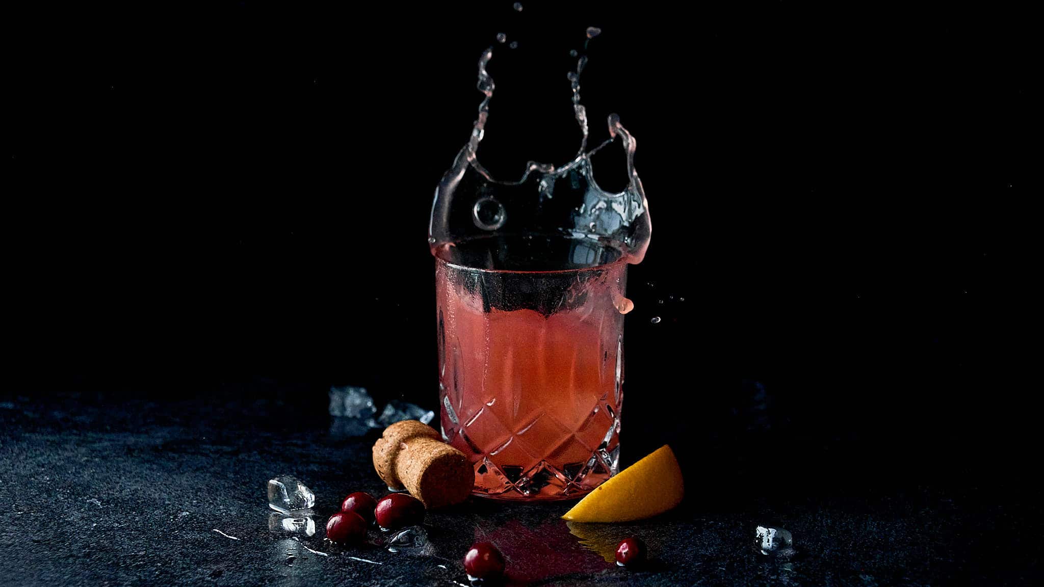 An old fashioned cocktail glass on a black background surrounded by an orange slice and cranberries as well as a champagne cork. The drink is splashing up and over the glass.