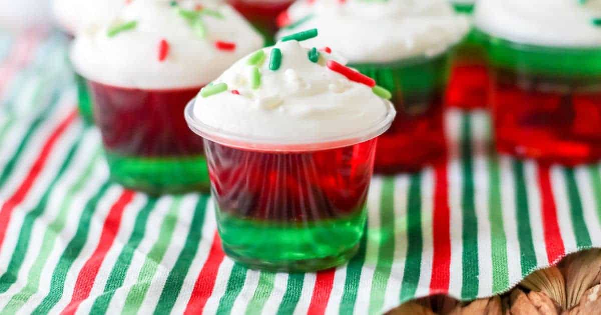 Several plastic cups filled with layered green and red jello shots, with whipped cream and sprinkles on top of each.