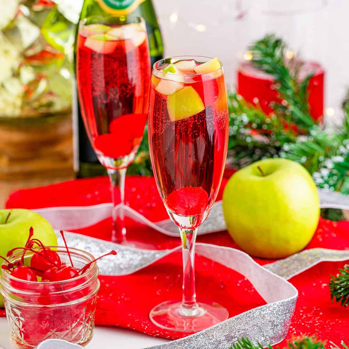 Two champagne glasses filled with bubbly red liquid, with cut up apples and red cherries for garnish.