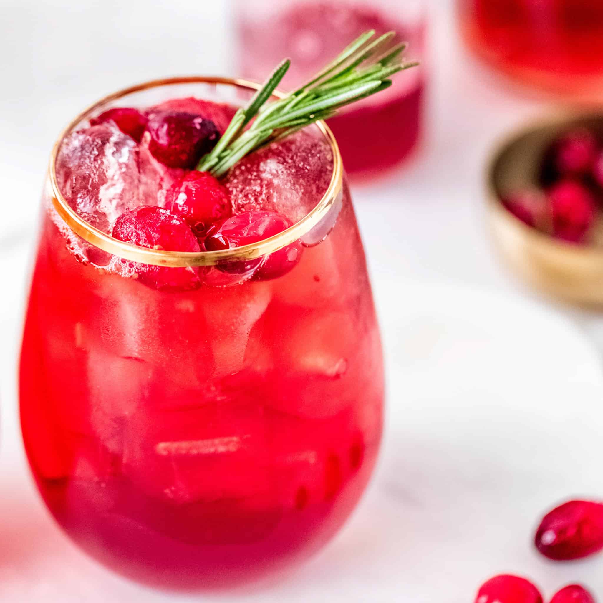 Gold rimmed stemless wine glass filled with a red cocktail and garnished with cranberries and rosemary sprig.